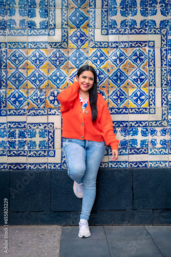 Full body of smiling young mexican woman looking at of camera while standing leaning with folded leg on wall of Sanborns restaurant in downtown Mexico City and touching hair in daylight photo