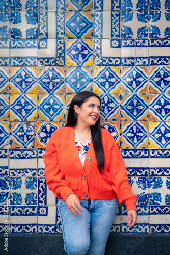 Full body of smiling young mexican woman looking at of camera while standing leaning with folded leg on wall of Sanborns restaurant in downtown Mexico City and touching hair in daylight photo