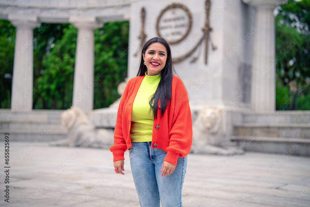 Fototapeta premium Smiling young Hispanic ethnic woman tourist smiling and looking away while standing in street in front of blurred Benito Juarez Hemicycle in Mexico city