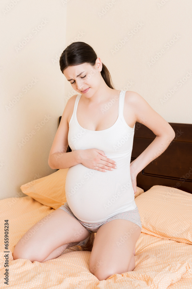 beautiful pregnant woman touching her tummy and keeping one hand on her back at home on bed beautiful pregnant woman touching her tummy and keeping one hand on her back at home on bed
