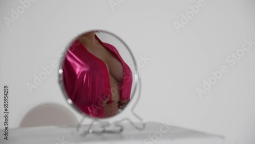 Small round mirror stands on white table in white room. Sexy caucasian plus size woman wearing pink robe on naked body reflects on mirror surface. Real time video. Abstract sensual background.
