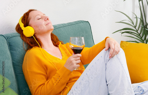 Woman with yellow headphones and a glass of wine sitting on the sofa at home and listening to music or podcast. music and relaxation