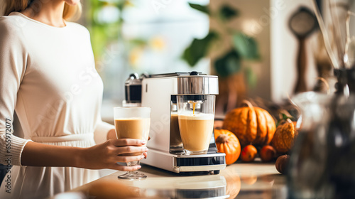 A young woman prepares pumpkin latte with milk on coffee machine in modern kitchen, daylight. Traditional autumn drink