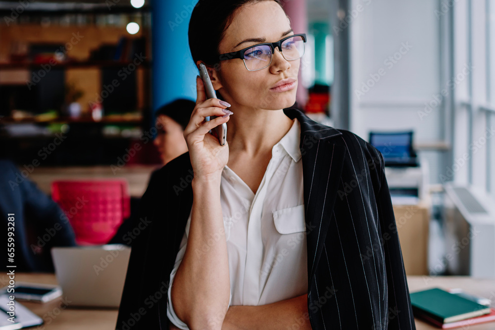 Serious female in eyewear looking away in office making mobile phone call for job Serious female in eyewear looking away in office making mobile phone call for job