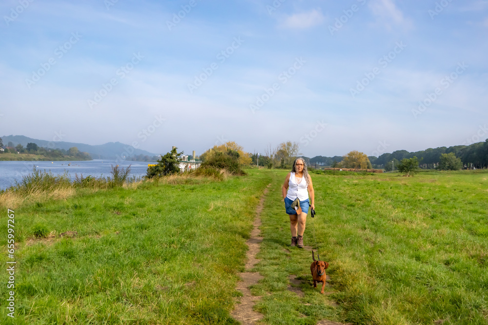 Plain next to Maas river with female hiker walking with her dog on trail, among green grass, hills and trees in blurred and misty background, sunny summer in Eijsden, South Limburg in the Netherlands