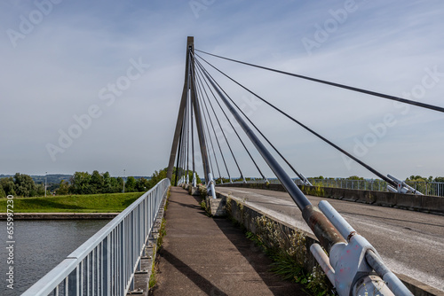 Pedestrian or cycling path parallel to rural road on Lanaye cable-stayed bridge, tower, pylon, huge turnbuckles and cables, trees, misty blue sky in background, wild plants, sunny day in Vise, Belgium