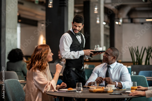 Young waiter serving two diverse business coworkers at the hotel cafe or bar photo