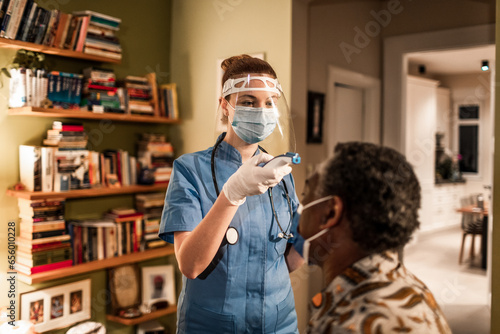 Young female caregiver fever screening her senior patient at their home photo