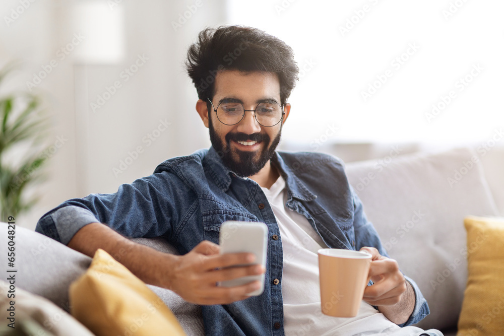 Domestic Leisure. Young Indian Man Using Smartphone And Drinking Coffee At Home Domestic Leisure. Young Indian Man Using Smartphone And Drinking Coffee At Home