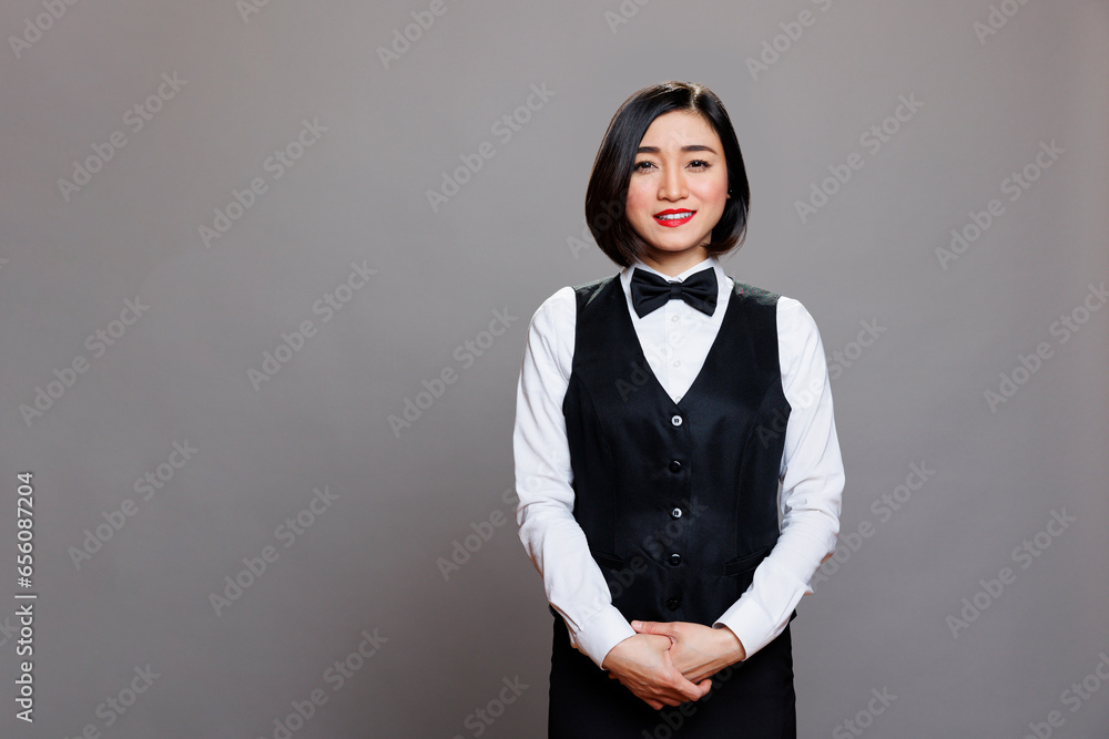 Smiling attractive asian waitress wearing uniform with bow tie standing and looking at camera. Restaurant professional receptionist posing for studio portrait on gray background Smiling attractive asian waitress wearing uniform with bow tie standing and looking at camera. Restaurant professional receptionist posing for studio portrait on gray background