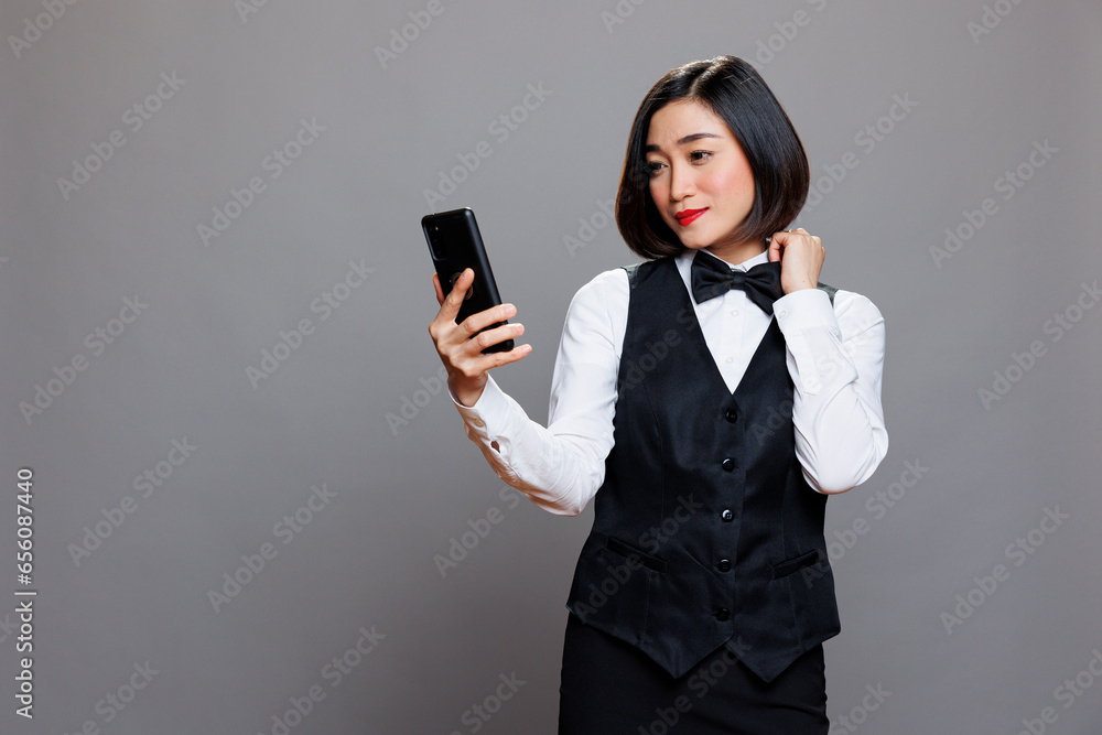 Smiling asian waitress looking at smartphone front camera while talking in videocall. Catering service attractive woman employee wearing uniform taking selfie on mobile phone