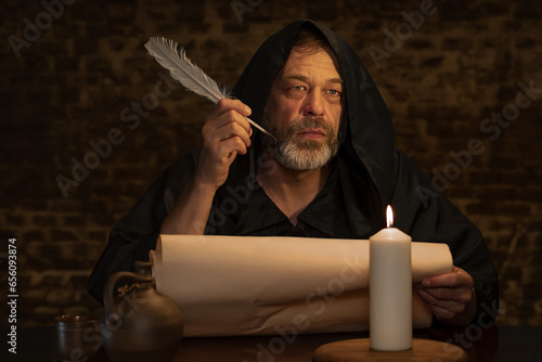 An old monk with a beard holds a quill in his hand over a parchment against a dark brick wall, dark tonality. photo