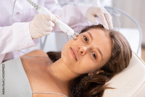 Obraz na plátně Young brown-haired woman receiving facial radiofrequency procedure stimulating collagen production resulting in tightened and firmed skin