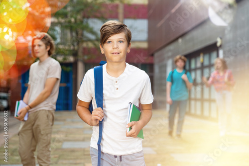 Young boy with backpack and copybooks in hand going home after classes in school. Senior students walking behind him.