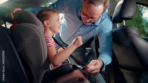 Dad puts her Kid girl in car seat safe car. Happy family. Father secures her kid in car seat using child safety belt. Family road trip Dad cares about her daughter safety. Child sits in car child seat