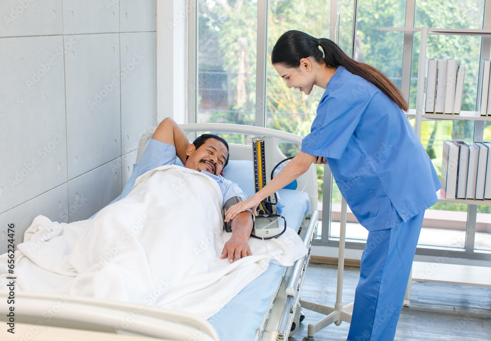 Asian professional experienced young female nurse in blue hospital uniform using wearing pressuring gauge around patient arm monitoring pressure laying lying down on bed under blanket in clinic ward Asian professional experienced young female nurse in blue hospital uniform using wearing pressuring gauge around patient arm monitoring pressure laying lying down on bed under blanket in clinic ward