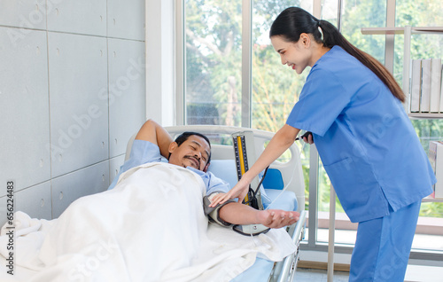 Asian professional experienced young female nurse in blue hospital uniform using wearing pressuring gauge around patient arm monitoring pressure laying lying down on bed under blanket in clinic ward photo