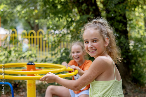 Two cute little blonde girls are spinning in a summer park on a swing. Leisure of schoolchildren during the holidays.