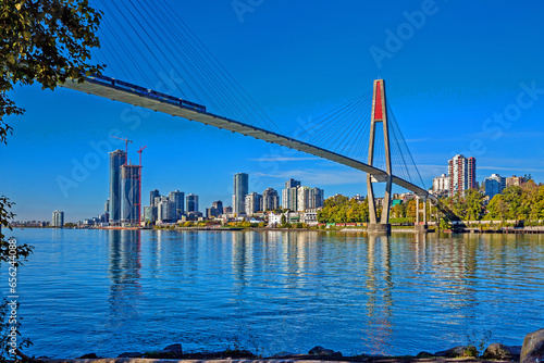 Skytrain bridge connecting Surrey and New Westminster over the Fraser River, New Westminster city center on the opposite bank, two high-rise buildings under construction, blue sky in the background