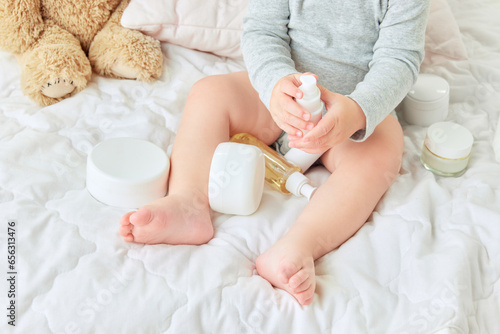 Cropped image of child legs, toddler sitting in bed with body moisturizing creams, lotions. Skincare products. Concept of childhood, kids cosmetology and natural cosmetics, body care