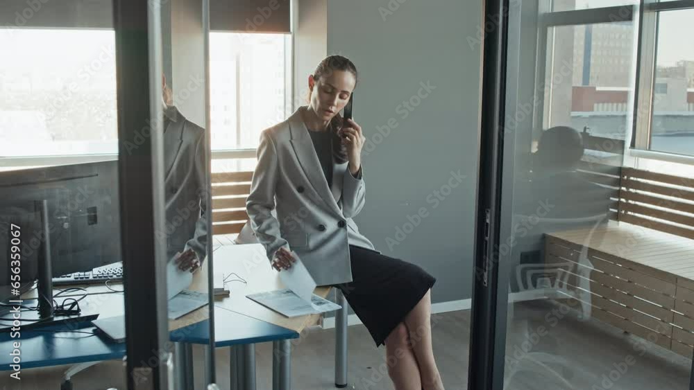 Through-doorway long slowmo shot of business woman sitting on table, speaking on phone, paging through and reading report Through-doorway long slowmo shot of business woman sitting on table, speaking on phone, paging through and reading report