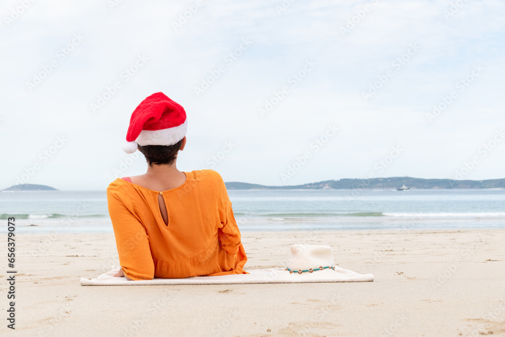 Woman on the beach sitting on her back with a red Christmas hat on and a white straw hat on the towel. Copy space.