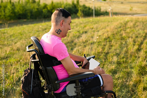 A disabled person with LGBT tattoo in a wheelchair works as a freelancer and uses a tablet while spending time in the park 