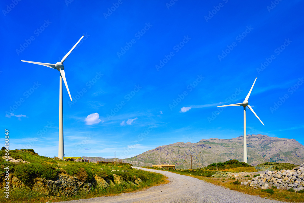 Two wind turbines by road on top of green hill on sunny day, Greece