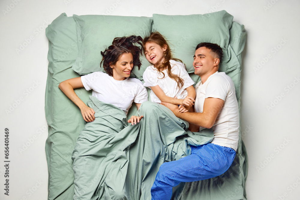 Top view portrait of cheerful people in bed. Cute child lying, having fun with parents in one bed with mint bedding. Enjoying being together. Top view portrait of cheerful people in bed. Cute child lying, having fun with parents in one bed with mint bedding. Enjoying being together.