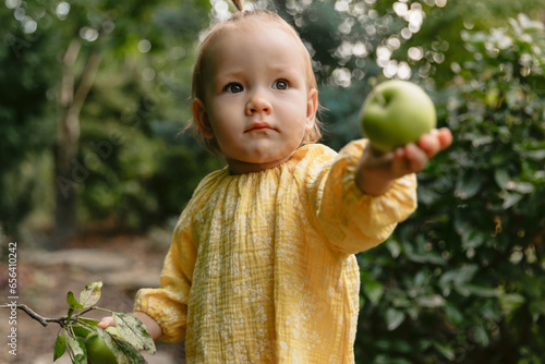 Portrait of cute girl gives an apple. Little girl in stylish dress in backyard garden.