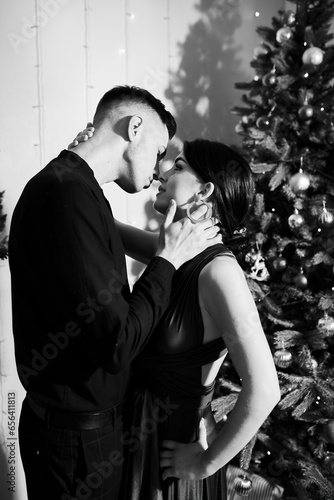 A young couple in love kiss on Christmas Eve by the Christmas tree with gifts at home in December. Black and white photo