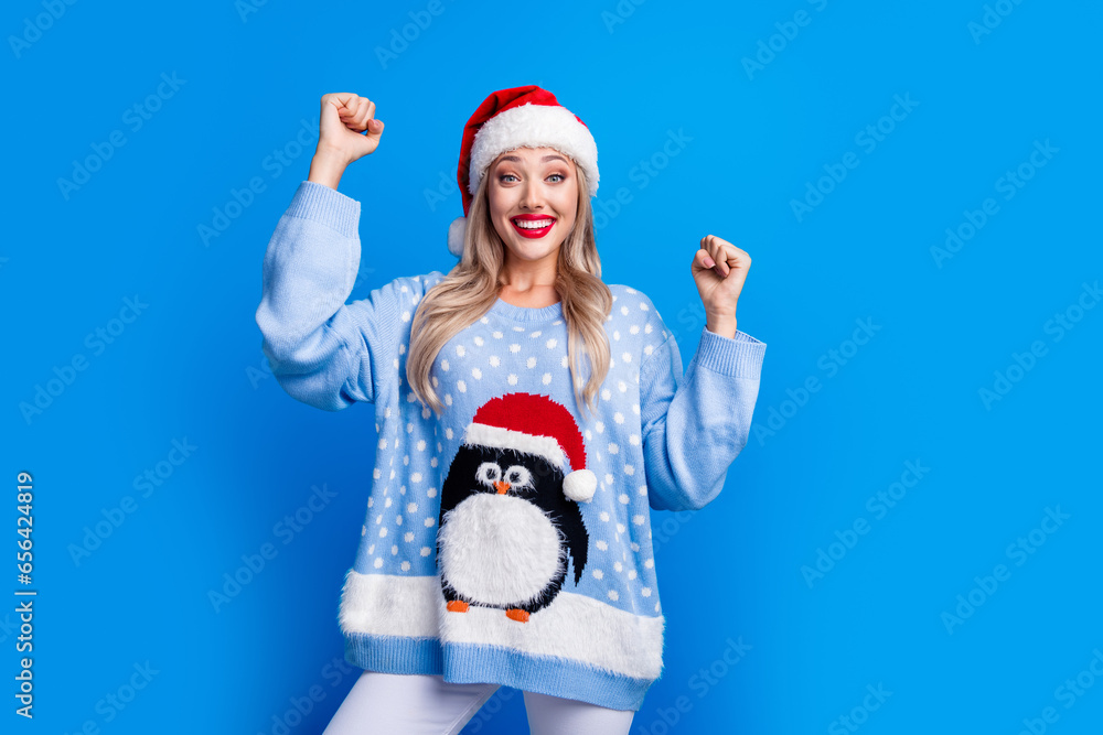 Obraz premium Photo portrait of attractive blonde young woman raise fists celebration wear penguin sweater x-mas hat isolated on blue color background
