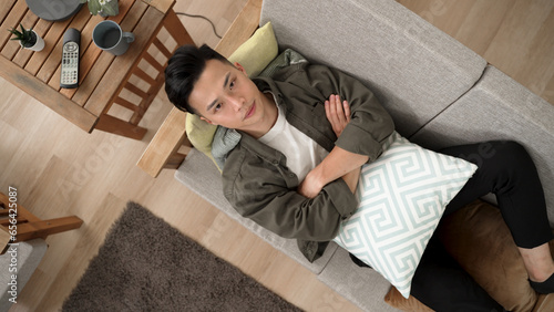 top view unhappy japanese male lying on uncomfortable couch with folded arms and pillow is looking upward in contemplation during quarantine.