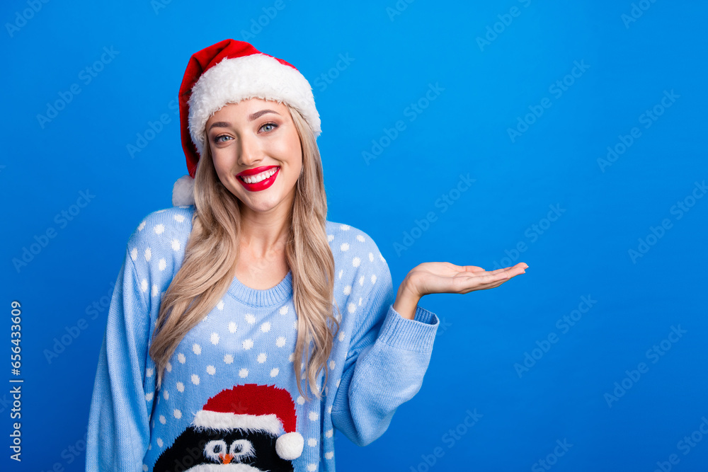 Obraz premium Photo portrait of lovely blonde young lady palm hold empty space wear penguin sweater x-mas hat isolated on blue color background