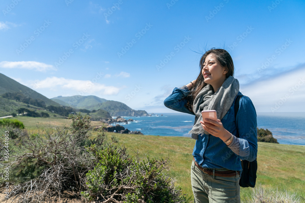 Naklejka premium happy asian chinese lady visitor enjoying California sun and breeze with phone in hand at beautiful big sur park in usa