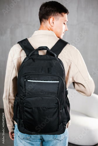 A black backpack is hanging on the back of a young guy in a light jacket