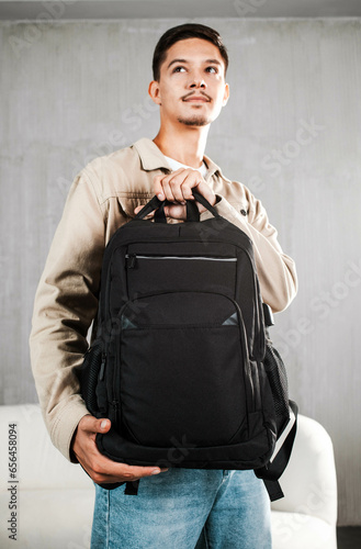 A handsome young man in a light jacket holds a black backpack by the upper strap
