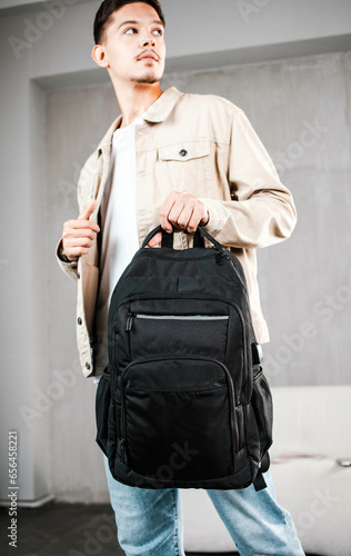 A handsome young man in a light jacket holds a black backpack by the upper strap