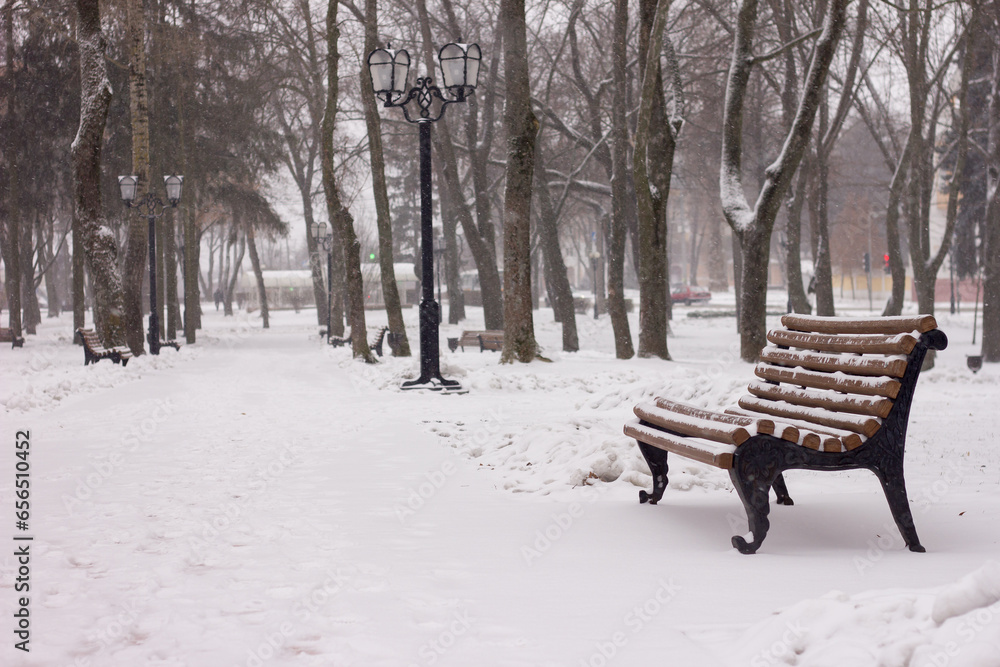 a bench sprinkled with snow and a street lamp near a path in a snowy park in winter