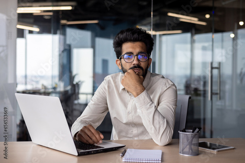 Portrait of a young Indian male student who is studying in the office at the table at the laptop, looking seriously and thoughtfully at the camera
