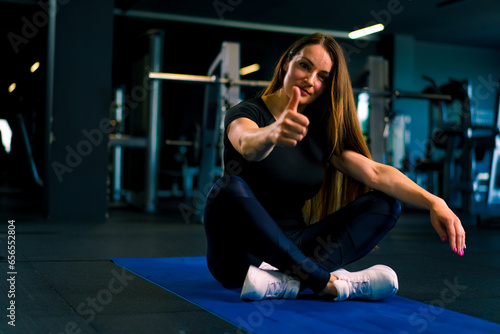young satisfied female fitness trainer showing super hand sign while sitting on sports mat in gym