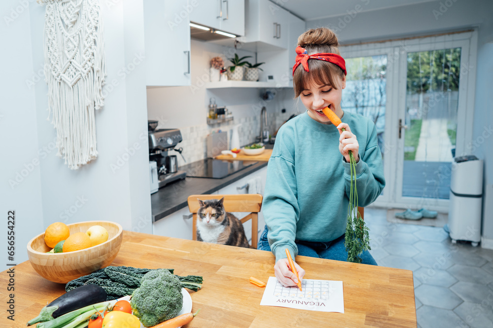 Young woman filling veganuary calendar while eating fresh carrot sitting on kitchen. Fresh vegetables and fruits on table. Vegan and vegetarian diet eating and cooking at home. Selective focus Young woman filling veganuary calendar while eating fresh carrot sitting on kitchen. Fresh vegetables and fruits on table. Vegan and vegetarian diet eating and cooking at home. Selective focus