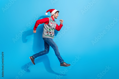 Full body photo of cute young man jump running fast hurry dressed x-mas print vest hat clothes isolated on blue color background