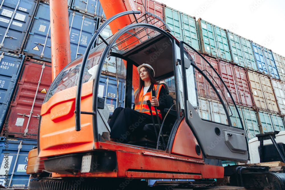 Caucasian woman in cabin wearing hard hat working as operator of loader crane machine in shipping containers terminal