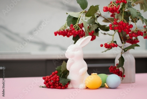 bouquet of mountain ash twigs in vase in home interior and white Easter bunnies on white table. In background is interior of white Scandinavian-style kitchen. Concept of home comfort