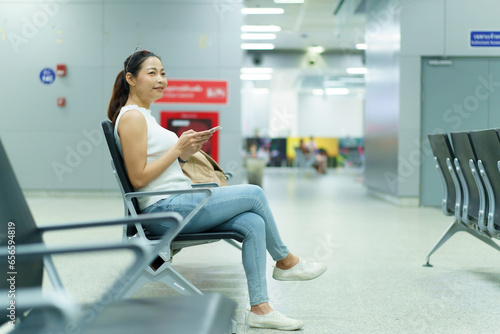 Happy beautiful Asian woman passenger sitting in airport departure terminal alone and sends a message or chats with her friends. Solo female traveler waiting for boarding at the gate in airport.
