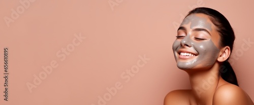Young woman enjoying a mud face mask treatment, with a joyful expression and eyes closed, isolated on a peachy hue backdrop with copy space