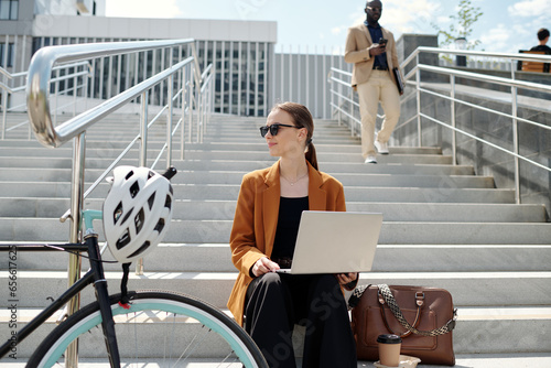 Young restful businesswoman with laptop on her knees looking aside while sitting on staircase in urban environment and enjoying short break photo