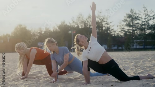 Three athletic beautiful girls doing yoga exercises on a sandy beach at dawn on a summer morning. The soft light of the sun in the background fills the scene. The wind shakes hair in slow motion
