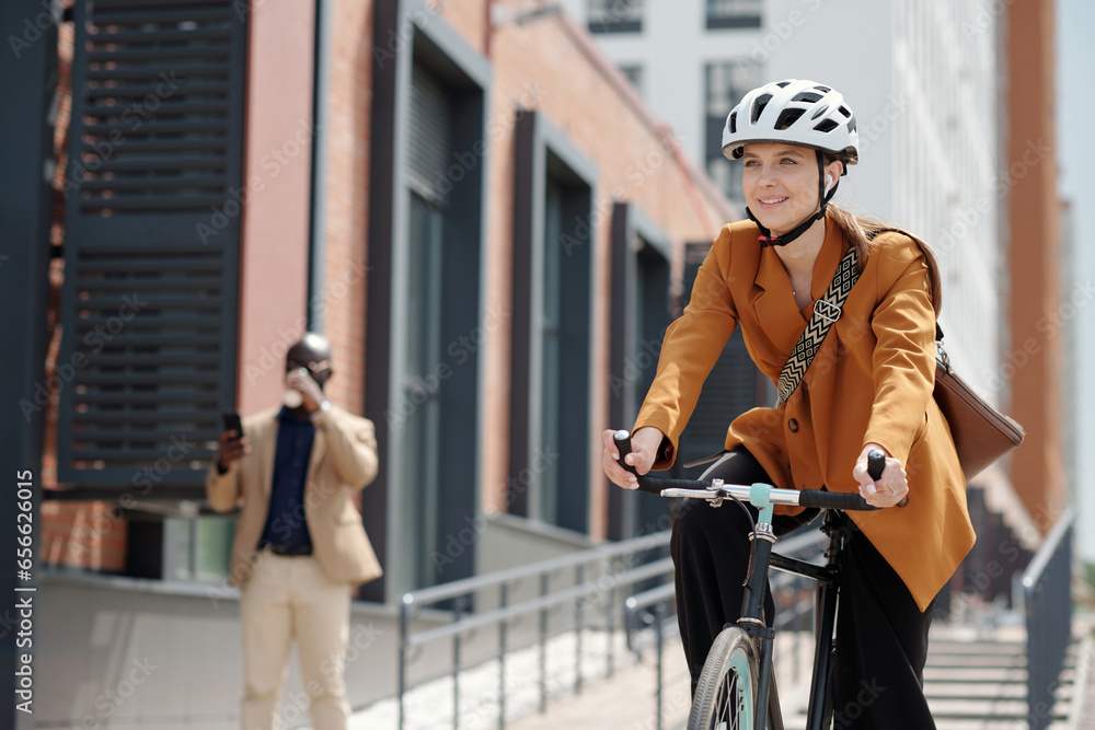 © pressmaster - Young cheerful female solopreneur in safety helmet and casualwear sitting on bicycle and riding home from work in urban environment
